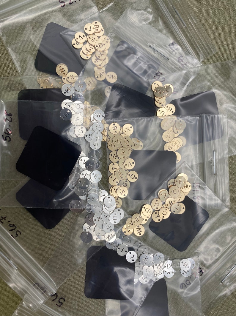May include: Three clear plastic bags filled with small round metal discs. The discs are silver, gold, and brass and have letters stamped on them. The bags are on a green surface.