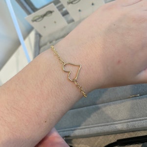 May include: A gold chain bracelet with a heart-shaped charm. The bracelet is on a person's wrist.