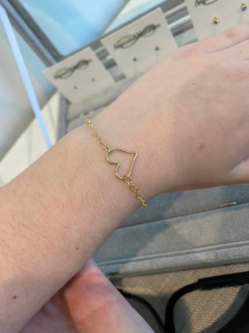May include: A gold chain bracelet with a heart-shaped charm. The bracelet is on a person's wrist.