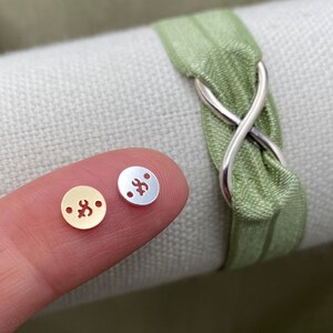 May include: Two small metal charms, one gold and one silver, with a decorative design. The charms are on a finger.