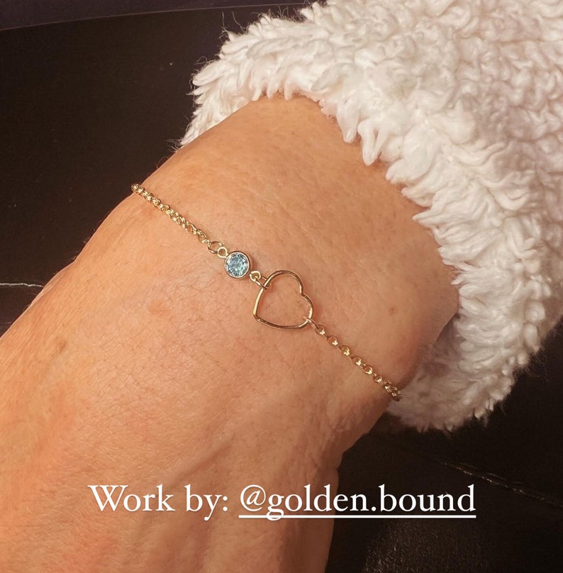 May include: A gold chain bracelet with a heart charm and a light blue gemstone. The bracelet is on a person's wrist. Work by: @golden.bound