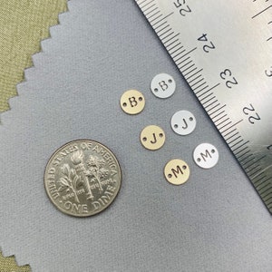May include: A close-up of five metal letter charms, each with a different letter. The letters are B, J, M, and two Bs. The charms are arranged in a row on a grey surface. A US dime is in the background for size comparison.