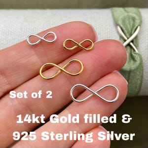 May include: A set of two infinity symbol charms, one in gold and one in silver, featuring a smooth, polished finish. The text on the image reads: Set of 2, 14kt Gold filled & 925 Sterling Silver. These small metal components are shaped like a figure-eight.