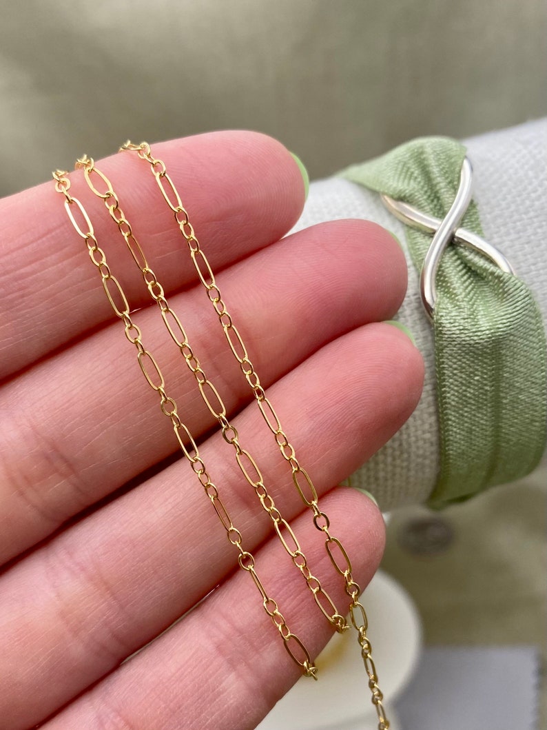 May include: Close-up of a delicate gold chain with elongated oval links. The chain is held in a hand with a light green painted fingernail. The background includes a light green fabric and a silver-colored metal accent.