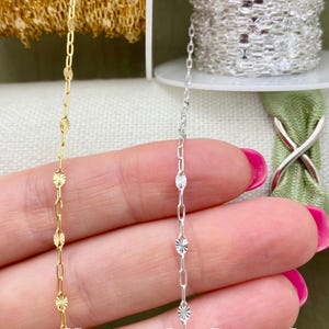 May include: Close-up of two delicate chains. One is gold-colored, featuring a repeating pattern of small, starburst-shaped charms linked by oval connectors. The other chain is silver-colored, with a similar design. Both chains are held against a soft background.
