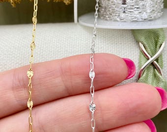 May include: Close-up of two delicate chains. One is gold-colored, featuring a repeating pattern of small, starburst-shaped charms linked by oval connectors. The other chain is silver-colored, with a similar design. Both chains are held against a soft background.