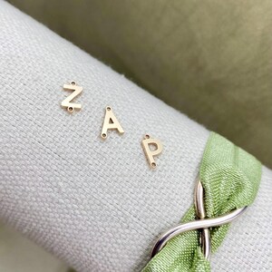 Pode incluir: Encantos de letras de metal dourado em forma das letras Z, A e P. Os encantos est&atilde;o sobre uma superf&iacute;cie branca com uma fita verde.