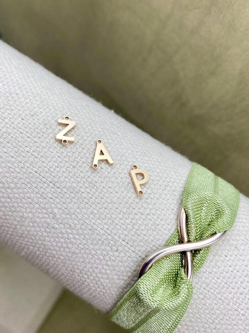 Pode incluir: Encantos de letras de metal dourado em forma das letras Z, A e P. Os encantos est&atilde;o sobre uma superf&iacute;cie branca com uma fita verde.