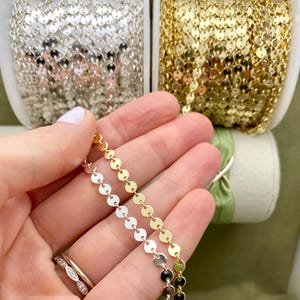 May include: Close-up of gold and silver disc chain jewellery. The chains are displayed on spools, with a hand holding a section of the gold and silver chain. The background includes spools of gold and silver chain.