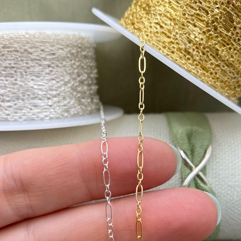 May include: Close-up of silver and gold chain. The silver chain is on the left, and the gold chain is on the right. Both chains have an oval and bar link design. The chains are on spools.
