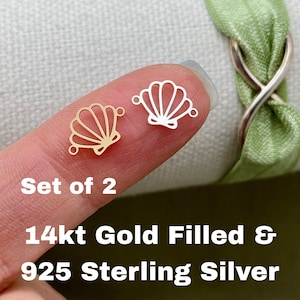 May include: Two seashell-shaped charms, one gold-colored and one silver-colored, are displayed. The charms are connected by small loops. The text "Set of 2" and "14kt Gold Filled & 925 Sterling Silver" are also visible.