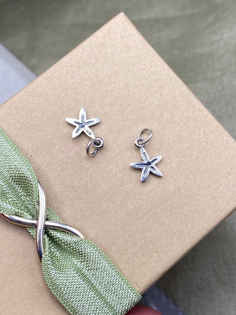 May include: Two silver starfish charms with a textured surface. The charms are on a brown surface with a green ribbon.