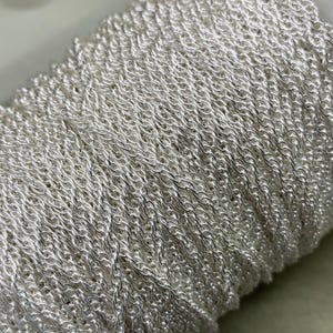 May include: A close-up of a silver chain wound onto a spool. The chain is made up of small, linked loops, creating a delicate and intricate design.