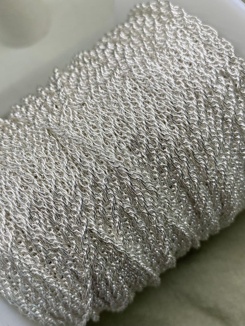 May include: A close-up of a silver chain wound onto a spool. The chain is made up of small, linked loops, creating a delicate and intricate design.