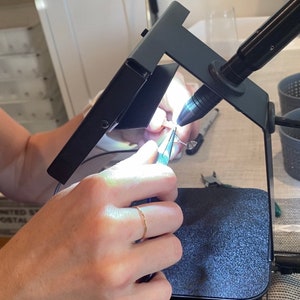 May include: A person uses tweezers to work on a small piece of jewelry under a magnifying lamp with a bright light. The jewelry is being worked on over a black mat.