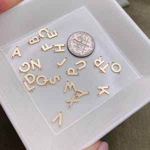 Pode incluir: Um prato de cer&acirc;mica branco cheio de pingentes de letras de metal dourado. As letras s&atilde;o de v&aacute;rios estilos e tamanhos. Uma moeda de dez centavos de d&oacute;lar americano est&aacute; inclu&iacute;da para escala.