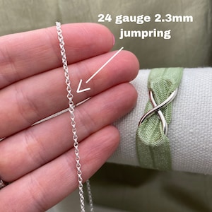 May include: A silver 24 gauge 2.3mm jump ring, shown in a hand holding the ring. The ring is a simple, round shape.
