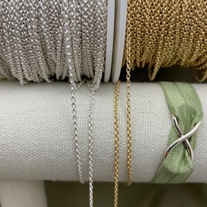 May include: Two spools of chain, one silver and one gold, are displayed on a white fabric surface. The chains are hanging down from the spools, showing their length and texture.