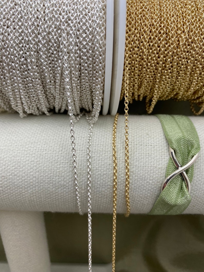 May include: Two spools of chain, one silver and one gold, are displayed on a white fabric surface. The chains are hanging down from the spools, showing their length and texture.