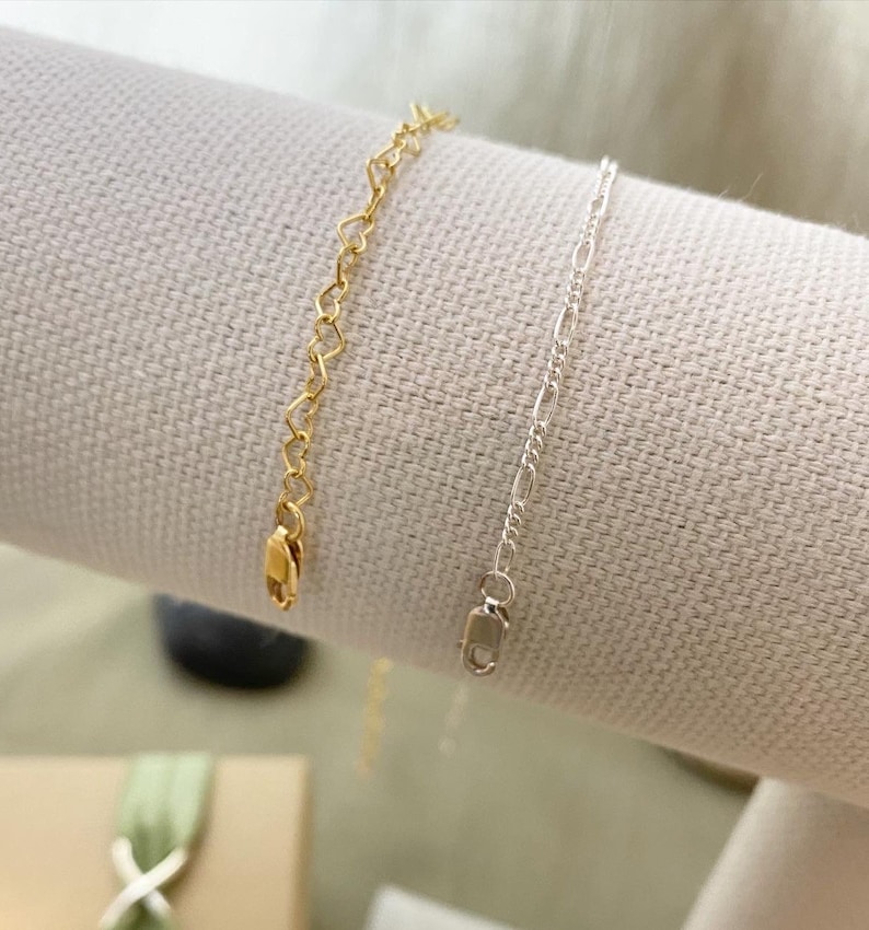 May include: Two delicate chain bracelets, one with a heart-shaped link in gold and the other with a simple chain link in silver.