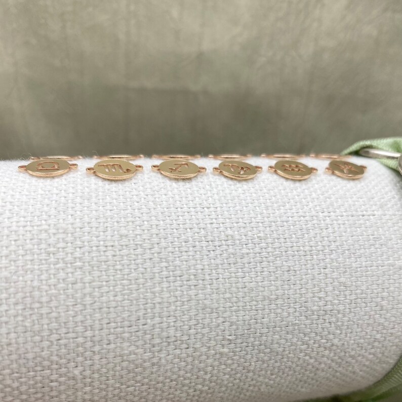 May include: A set of gold-toned zodiac charms, each featuring a different astrological sign. The charms are arranged in a row on a white fabric background.