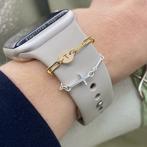 May include: A white Apple Watch with a gold chain bracelet and a silver cross charm bracelet. The chain bracelet has a gold disc charm with the letter "T" engraved on it. The watch band is a white silicone band. The watch is on a person's wrist.