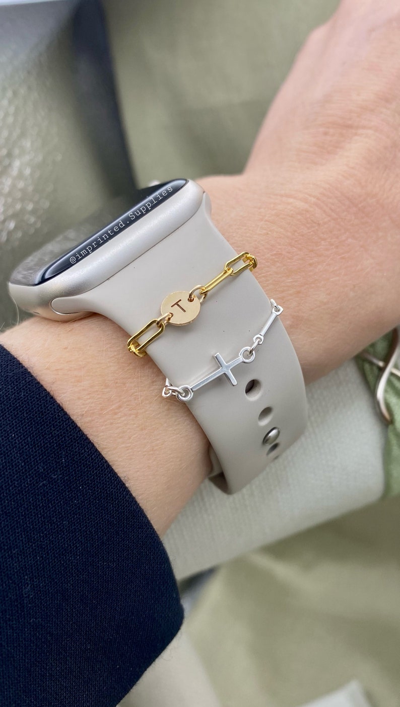 May include: A white Apple Watch with a gold chain bracelet and a silver cross charm bracelet. The chain bracelet has a gold disc charm with the letter "T" engraved on it. The watch band is a white silicone band. The watch is on a person's wrist.