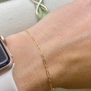 May include: A delicate gold-colored bracelet featuring an oval link chain. The bracelet is worn on a wrist, with a gold-colored watch and a green fabric accessory in the background. The bracelet is a simple, elegant piece of jewelry.