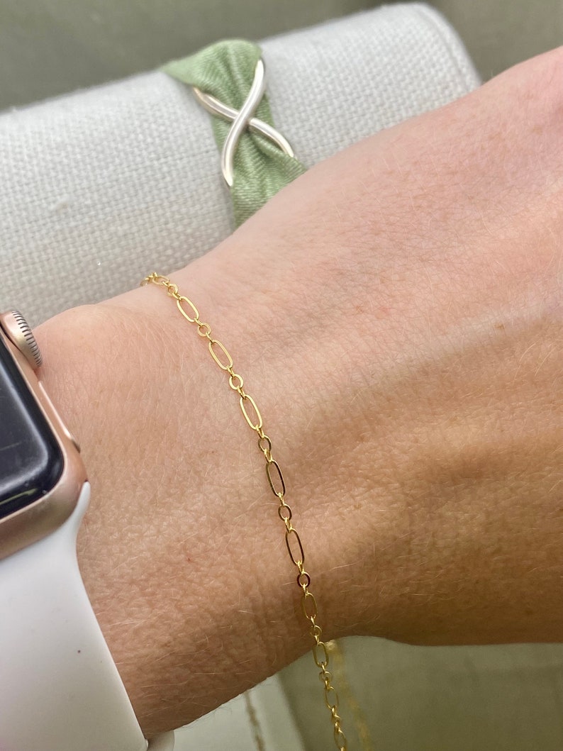 May include: A delicate gold-colored bracelet featuring an oval link chain. The bracelet is worn on a wrist, with a gold-colored watch and a green fabric accessory in the background. The bracelet is a simple, elegant piece of jewelry.