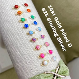 May include: A variety of opal connector charms in 14kt gold filled and 925 sterling silver. The charms are displayed on a white fabric surface. The charms are available in a variety of colors, including red, orange, yellow, green, blue, pink, and white.