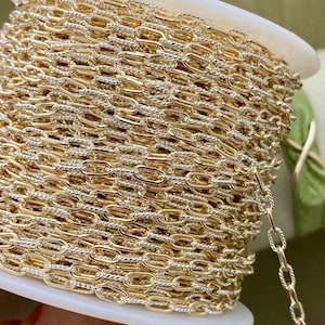 May include: A spool of gold-colored chain with a silver-colored twisted chain design. The chain is wound around a white plastic spool. The chain is likely used for jewelry making or other craft projects.