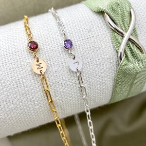 May include: Two gold chain bracelets with a small gemstone and a letter charm. The bracelet on the left has a red gemstone and the letter "M" charm. The bracelet on the right has a purple gemstone and the letter "J" charm.