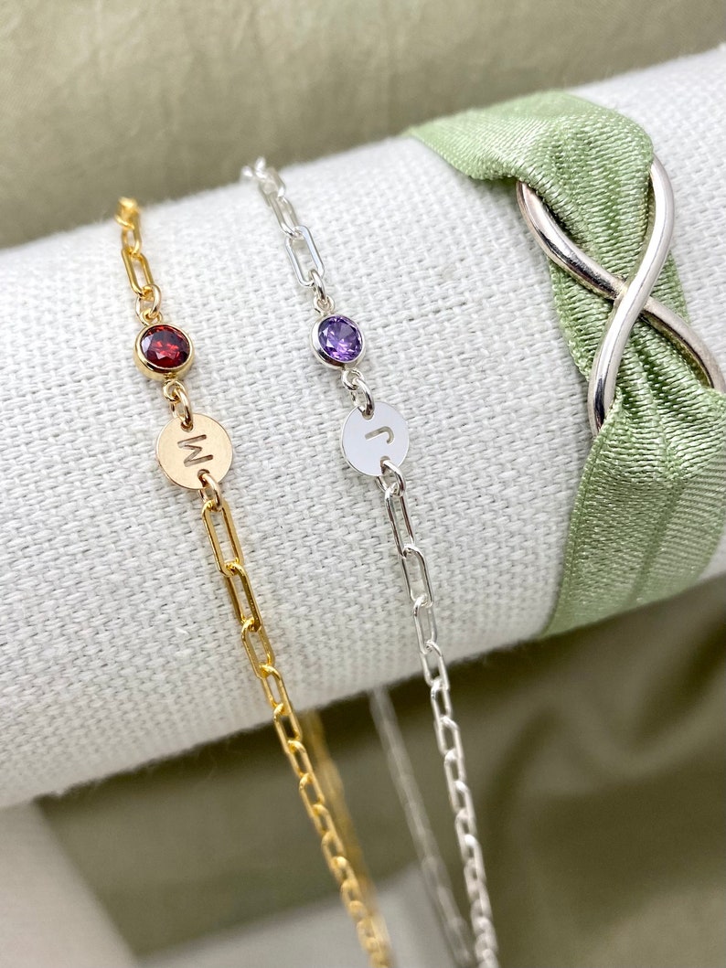 May include: Two gold chain bracelets with a small gemstone and a letter charm. The bracelet on the left has a red gemstone and the letter "M" charm. The bracelet on the right has a purple gemstone and the letter "J" charm.