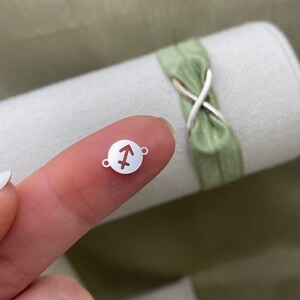 May include: A silver charm with a Sagittarius zodiac symbol cut out. The charm is round and has two small holes for attaching to a bracelet or necklace.
