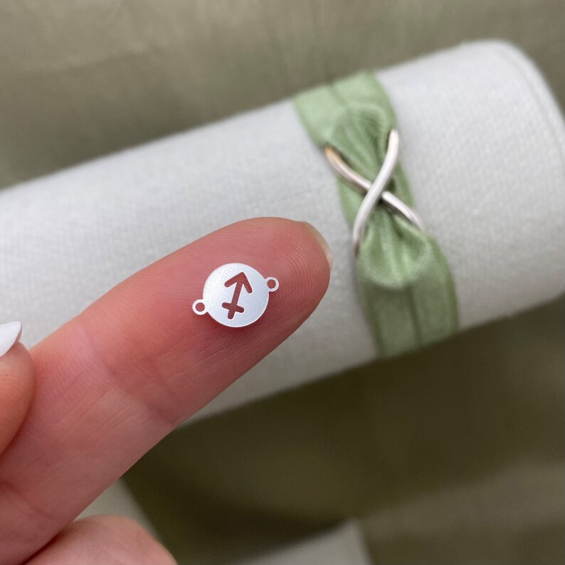 May include: A silver charm with a Sagittarius zodiac symbol cut out. The charm is round and has two small holes for attaching to a bracelet or necklace.
