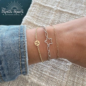 May include: Three gold chain bracelets on a wrist. The top bracelet has a star charm, the middle bracelet has an Om symbol charm, and the bottom bracelet is a simple chain.
