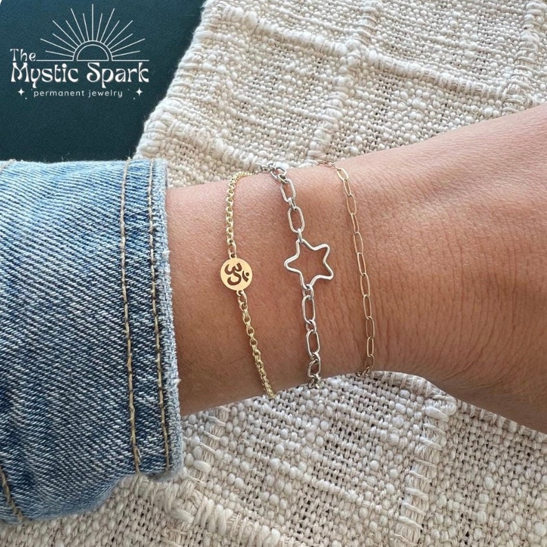May include: Three gold chain bracelets on a wrist. The top bracelet has a star charm, the middle bracelet has an Om symbol charm, and the bottom bracelet is a simple chain.