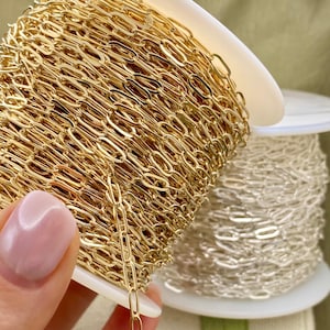 May include: Close-up of gold-colored chain links on a white spool. The chain is tightly wound, with a second spool of silver-colored chain visible in the background. The chain is likely used for jewelry making or crafting.