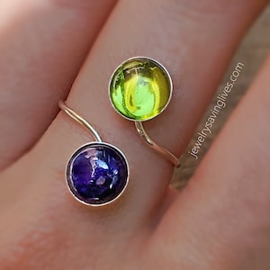 May include: A silver ring with two round glass stones. The larger stone is green and yellow, and the smaller stone is purple. The ring is on a finger.