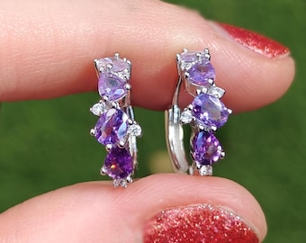 Può includere: Un paio di orecchini a cerchio in argento, ciascuno ornato da un gruppo di pietre preziose viola e lavanda. Gli orecchini presentano un mix di pietre ovali e a forma di pera, impreziosite da piccoli cristalli trasparenti.