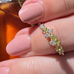 May include: A delicate gold ring featuring a cluster of small green and white gemstones. The ring is worn on a finger with pink nail polish.
