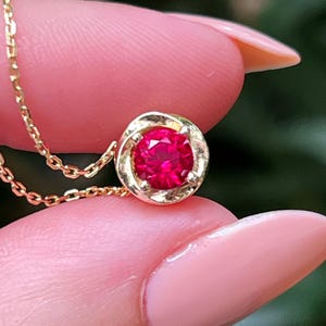 May include: A gold necklace with a bright red gemstone pendant. The round gemstone is set in a gold bezel, and the necklace has a fine chain. The jewellery is displayed against a blurred green background.