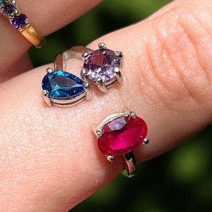 Puede incluir: Una colección de anillos con piedras preciosas de varios colores. Un anillo presenta un arcoíris de piedras, mientras que otros muestran gemas azules, moradas y rojas. Los anillos están engarzados en bandas de plata u oro, creando una exhibición vibrante de joyería.