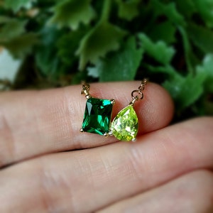 May include: A gold necklace featuring two gemstones: a rectangular emerald-cut green stone and a pear-shaped light green stone. The necklace is displayed against a blurred green background, highlighting the vibrant colors of the gems.