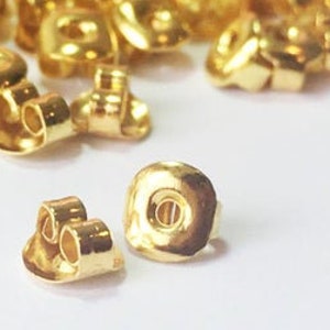 May include: A close-up of a pile of gold-colored earring backs. The backs are made of metal and have a small hole in the center. Some of the backs are in the shape of a butterfly.