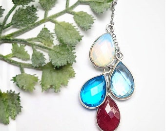 Puede incluir: Un collar de cadena de plata con un grupo de cuatro piedras preciosas en forma de lágrima. Las piedras preciosas son blanco iridiscente, azul claro, azul oscuro y rojo.