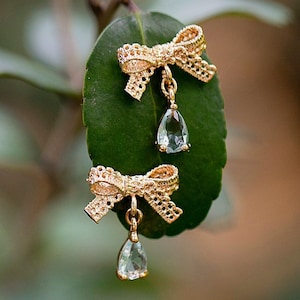 May include: A pair of gold earrings with a delicate bow design and a teardrop-shaped clear gemstone hanging from each bow.