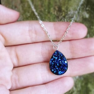 May include: A silver chain necklace with a teardrop-shaped pendant. The pendant is made of blue glitter embedded in a clear resin.