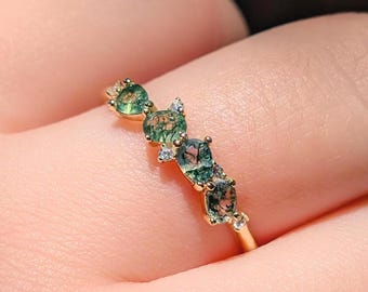 May include: A gold ring featuring four oval-shaped moss agate stones with green inclusions, accented by small clear round stones set between the larger gems. The band is thin and metallic, showcasing a delicate arrangement of stones.