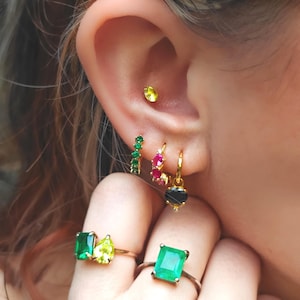 Könnte beinhalten: Eine Auswahl an goldfarbenem Schmuck, darunter Ohrringe und Ringe, mit verschiedenen farbigen Edelsteinen. Die Ohrringe umfassen einen kleinen gelben Ohrstecker, einen grünen und roten Stab sowie einen goldenen Creolen mit einem schwarzen Stein. Ringe zeigen Smaragd- und gelb-grüne Steine.